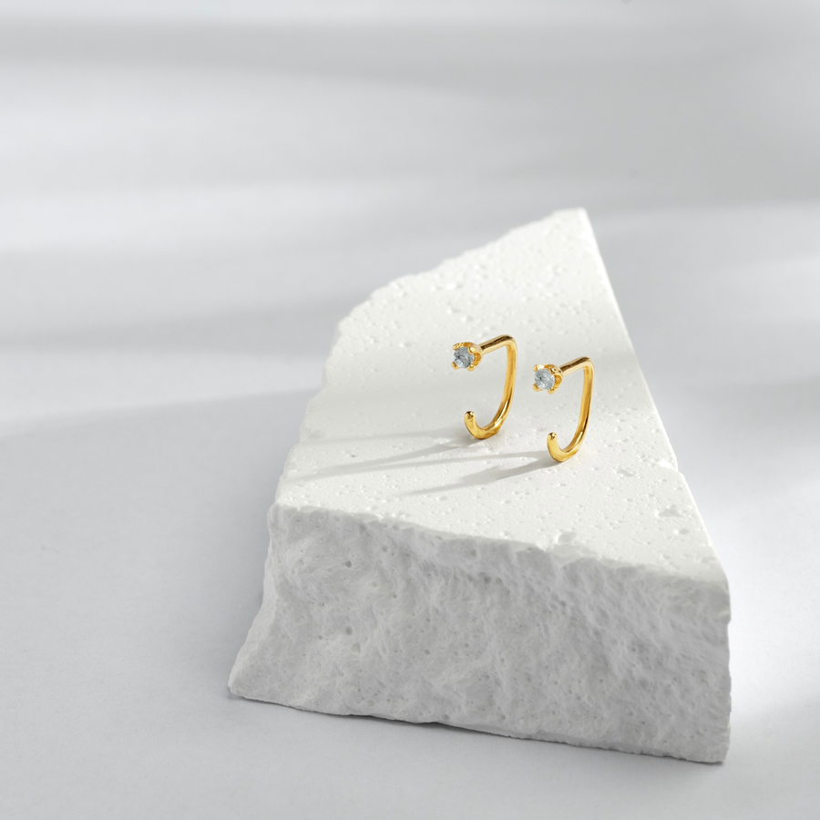 Gold Stay Close Aquamarine Hoops on a white marble stand against a light gray background